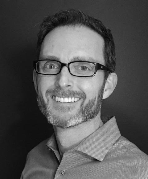 Black-and-white headshot of a smiling man wearing glasses and a collared shirt, with a short beard, photographed against a plain dark background.