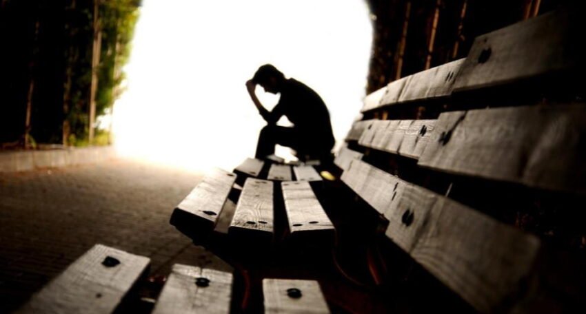 Silhouette of a person sitting alone on a wooden bench inside a tunnel, head in hands, with bright light at the end symbolizing distress or emotional struggle.