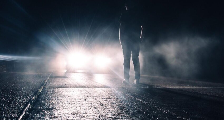 A person standing on a dark, wet road at night facing a car with bright headlights shining through fog.