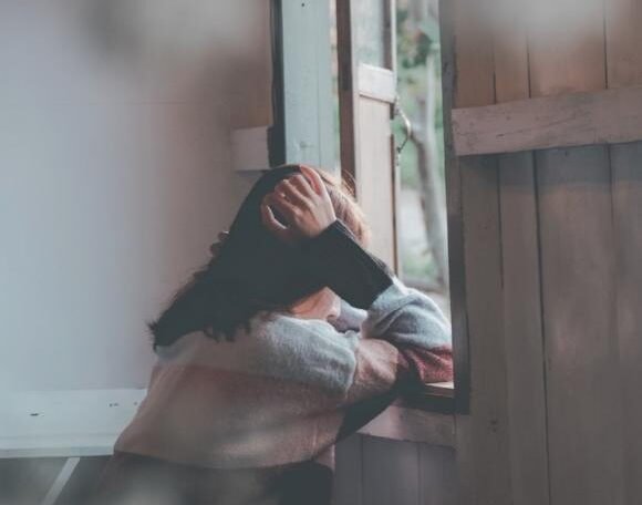 Person sitting by an open window with head in hands, expressing sadness or deep thought in a quiet room