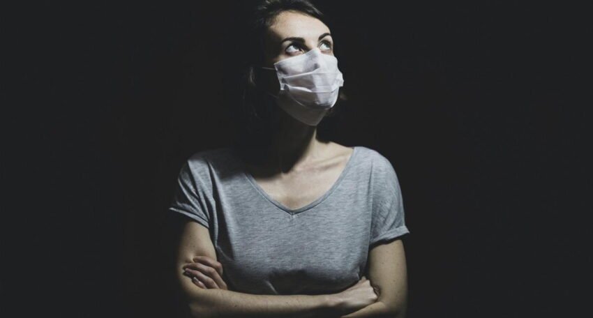 Woman wearing a face mask standing with crossed arms against a dark background, looking upward
