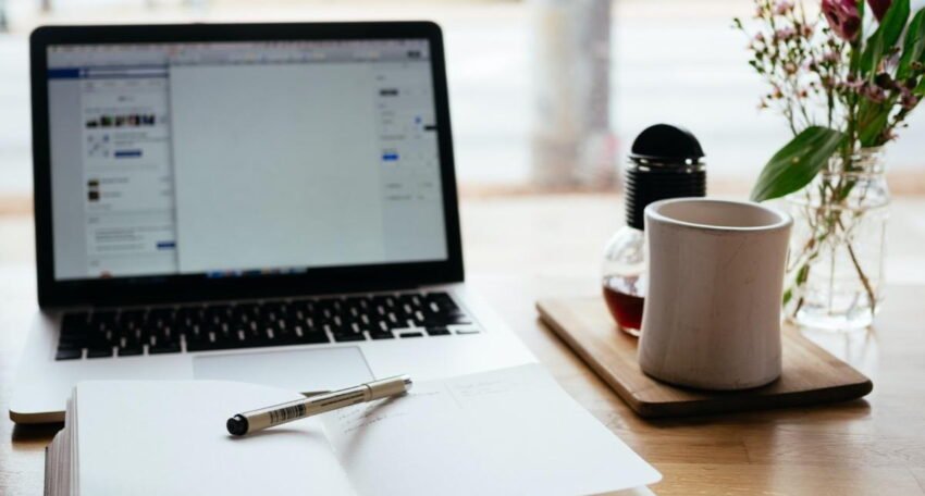 Open notebook with a pen on a wooden desk beside a laptop and coffee cup in a bright workspace