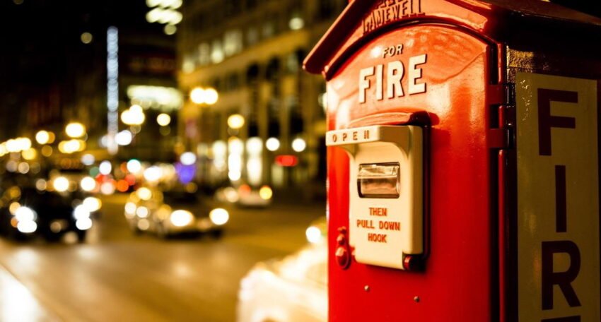 Red fire alarm call box on a city street at night, with blurred car headlights and illuminated buildings in the background.